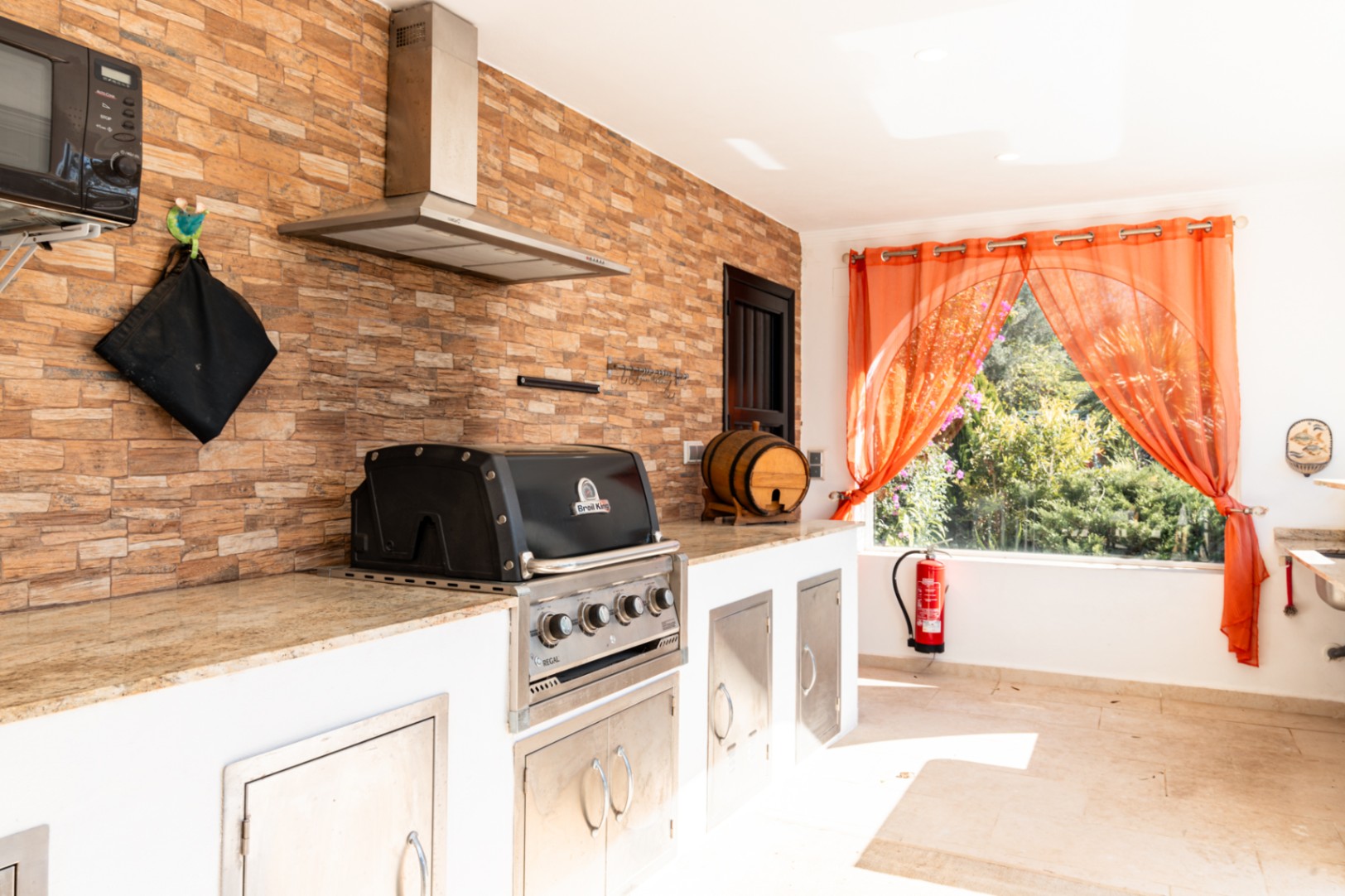 Cuisine d'été équipée d'un barbecue Broil King encastré, d'un plan de travail en granit et d'un mur en pierre naturelle avec hotte aspirante en inox.