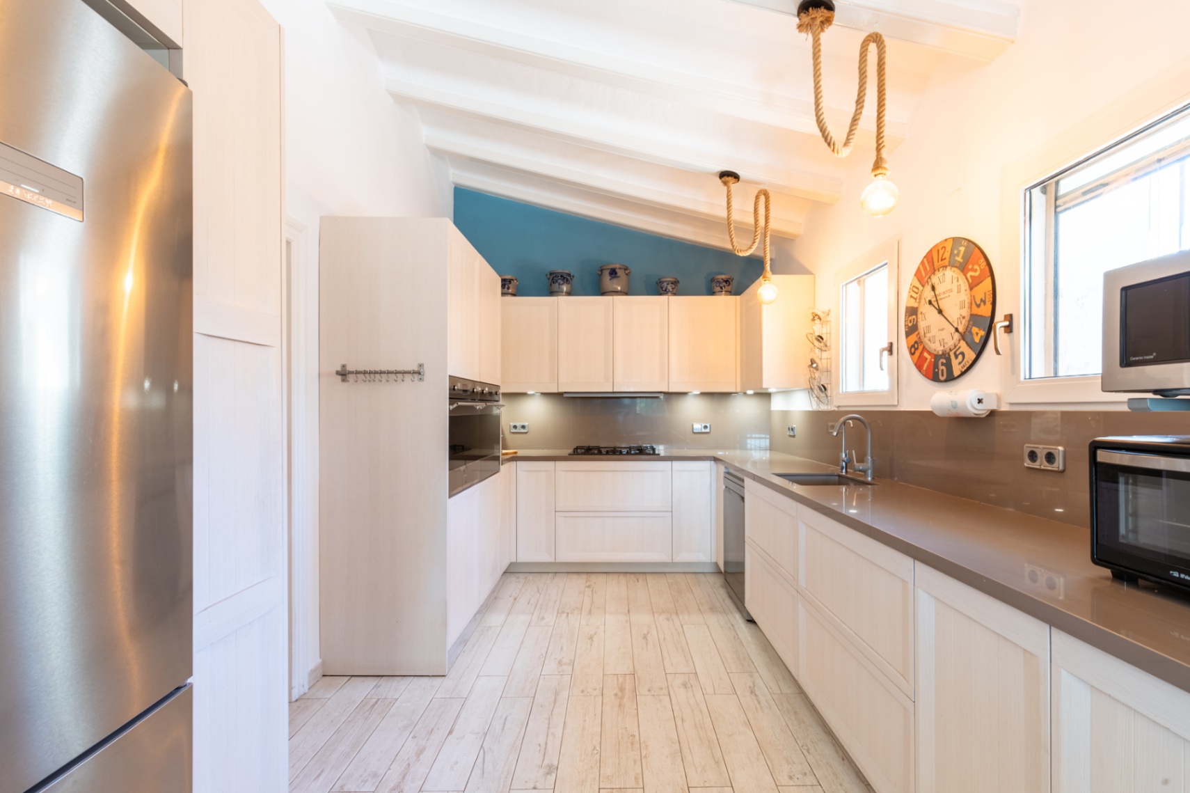 Cuisine équipée avec plans de travail en pierre, électroménager en inox et plafond incliné à poutres. Elle dispose de luminaires en corde et d'un mur d'accent bleu.