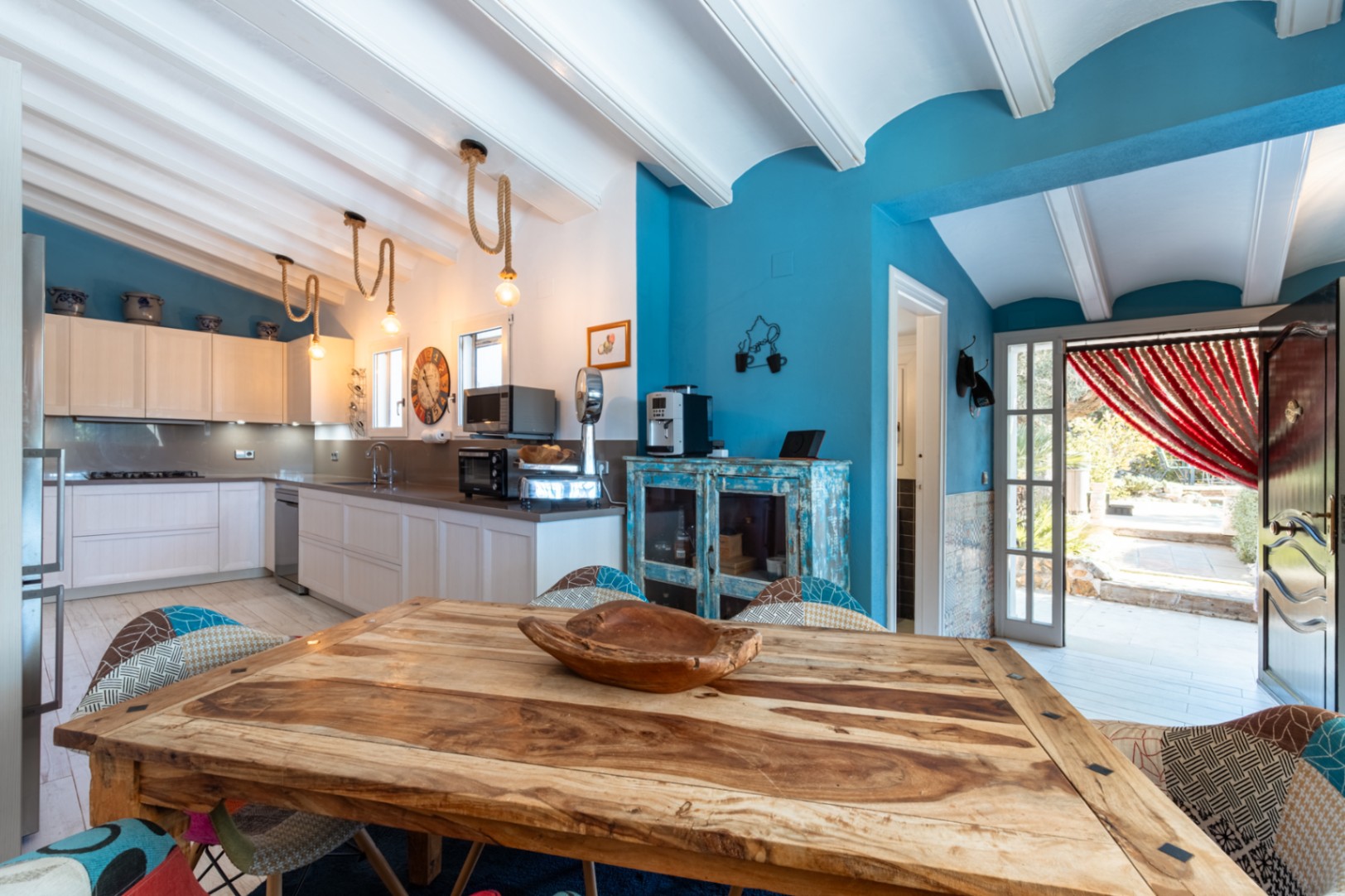 Cuisine moderne avec plafonds à poutres blanches, murs bleu vif, table en bois rustique et suspensions en corde. Équipée d'appareils haut de gamme et accès extérieur.