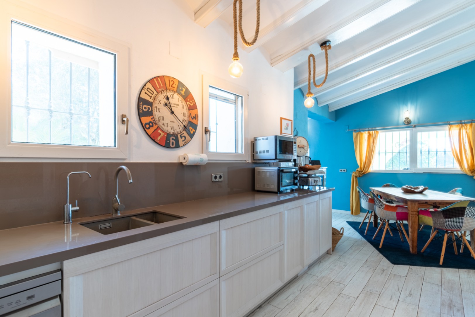 Cuisine moderne avec plan de travail en quartz, ouverte sur un espace repas lumineux aux murs bleus, poutres blanches apparentes et luminaires suspendus en corde.