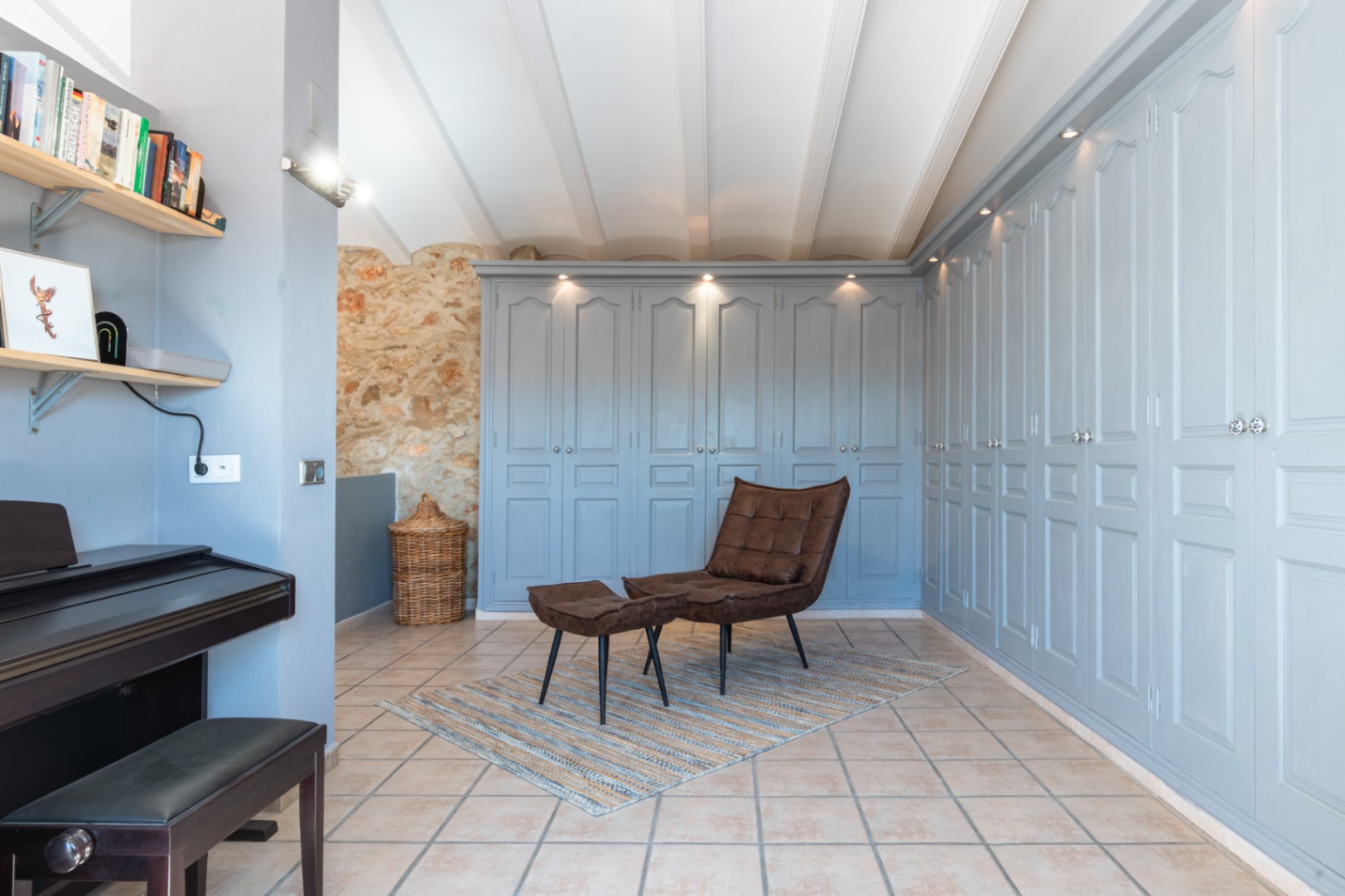 Espace de transition avec de grands placards intégrés bleu pastel, mur en pierre naturelle, plafond à poutres blanches et un coin détente avec fauteuil en cuir et piano.
