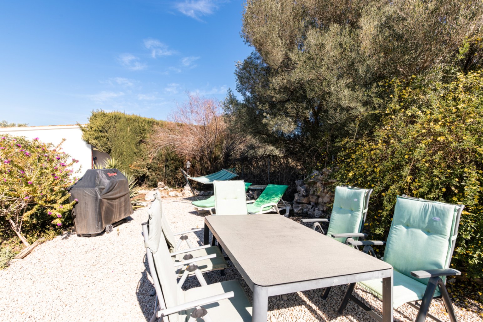 Espace dînatoire extérieur sur gravier avec table rectangulaire, chaises rembourrées, barbecue protégé et hamac au cœur d'un jardin arboré.