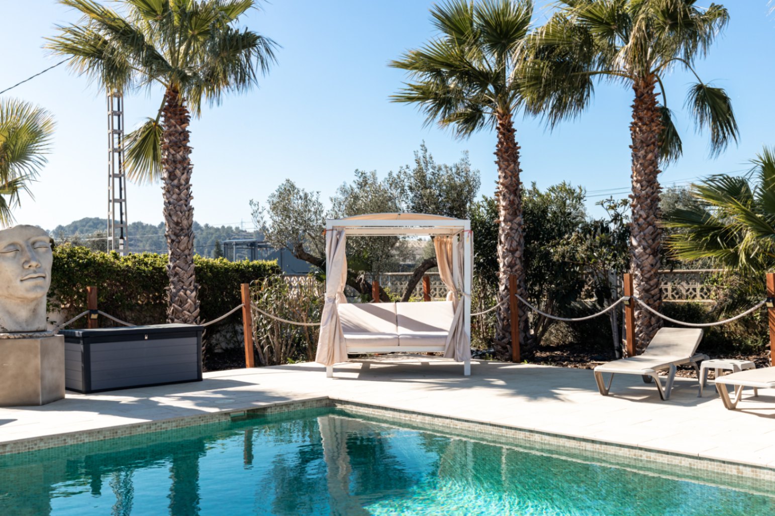Espace piscine avec terrasse en pierre, lit balinais blanc à rideaux, bains de soleil et palmiers matures entourant le bassin.