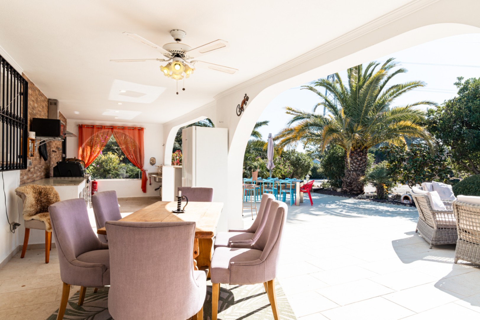 Grande naya couverte avec coin repas, ventilateur de plafond et cuisine d'été. L'espace s'ouvre sur un jardin ensoleillé agrémenté de palmiers et d'un salon extérieur.