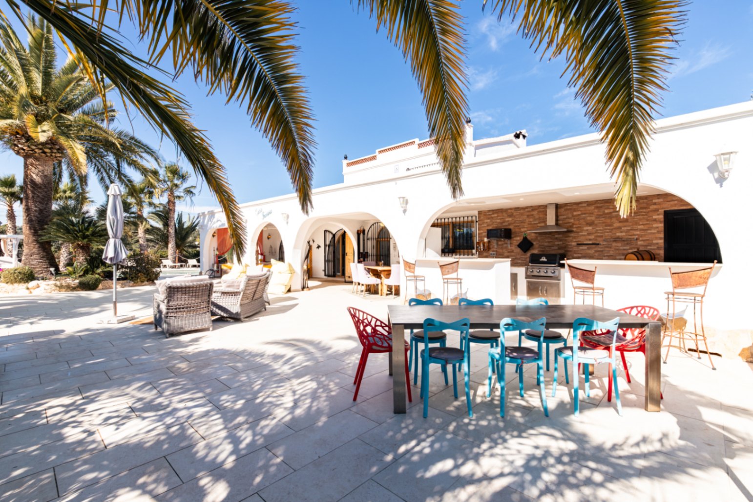 Grande terrasse extérieure avec naya voûtée, cuisine d'été équipée d'un barbecue Broil King et espace dînatoire entouré de palmiers matures.