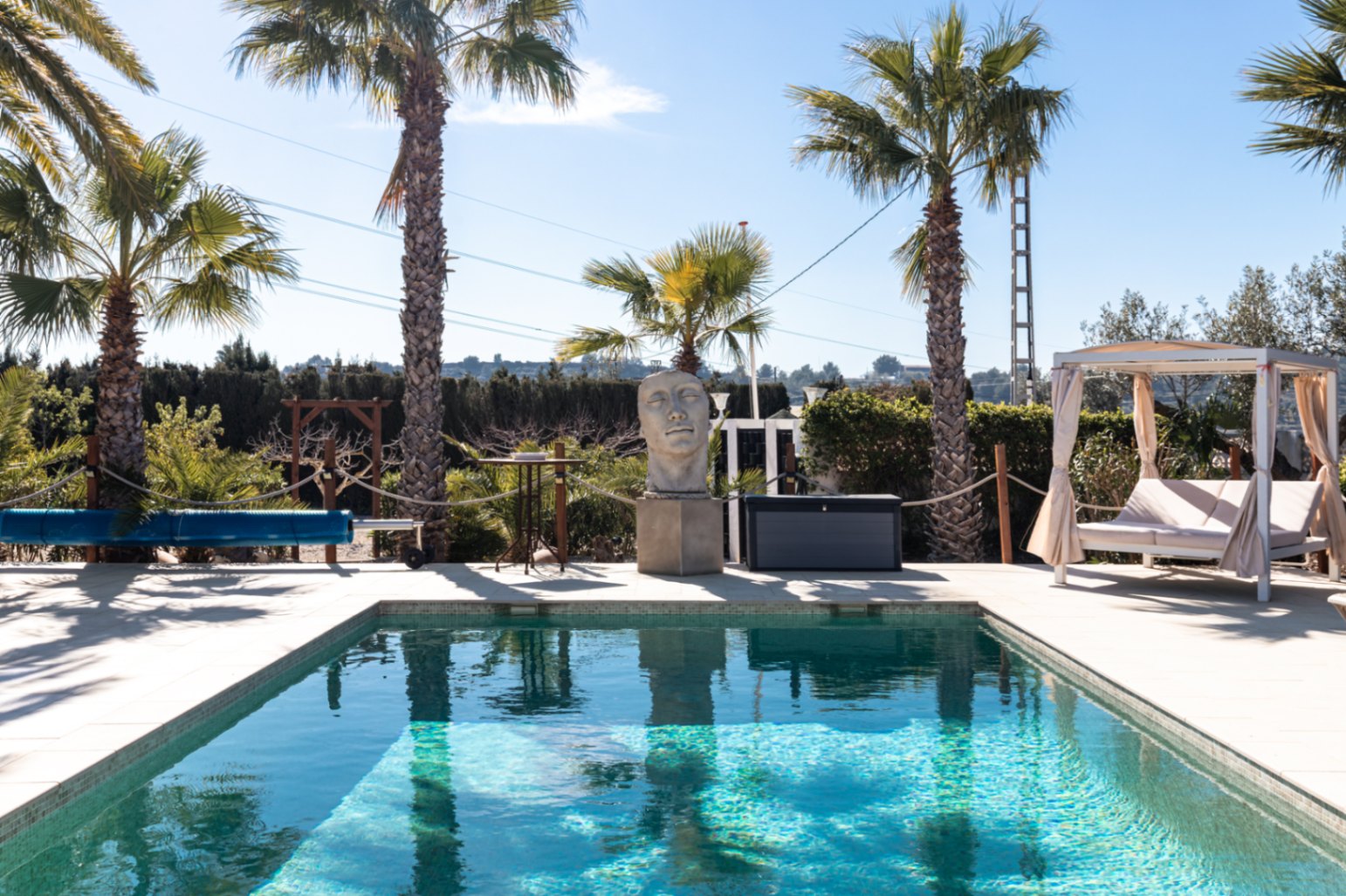 Piscine de 9,5m x 5m avec terrasse en pierre, sculpture décorative, lit balinais et couverture de protection, entourée de palmiers sous un ciel dégagé.