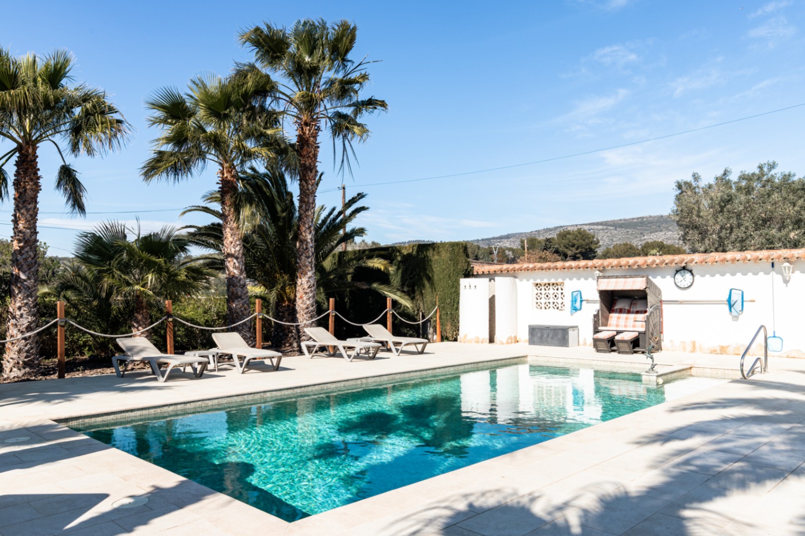 Piscine de 9,5m x 5m entourée d'une terrasse en pierre claire, avec transats, palmiers et un fauteuil de plage traditionnel sous un ciel azur méditerranéen.