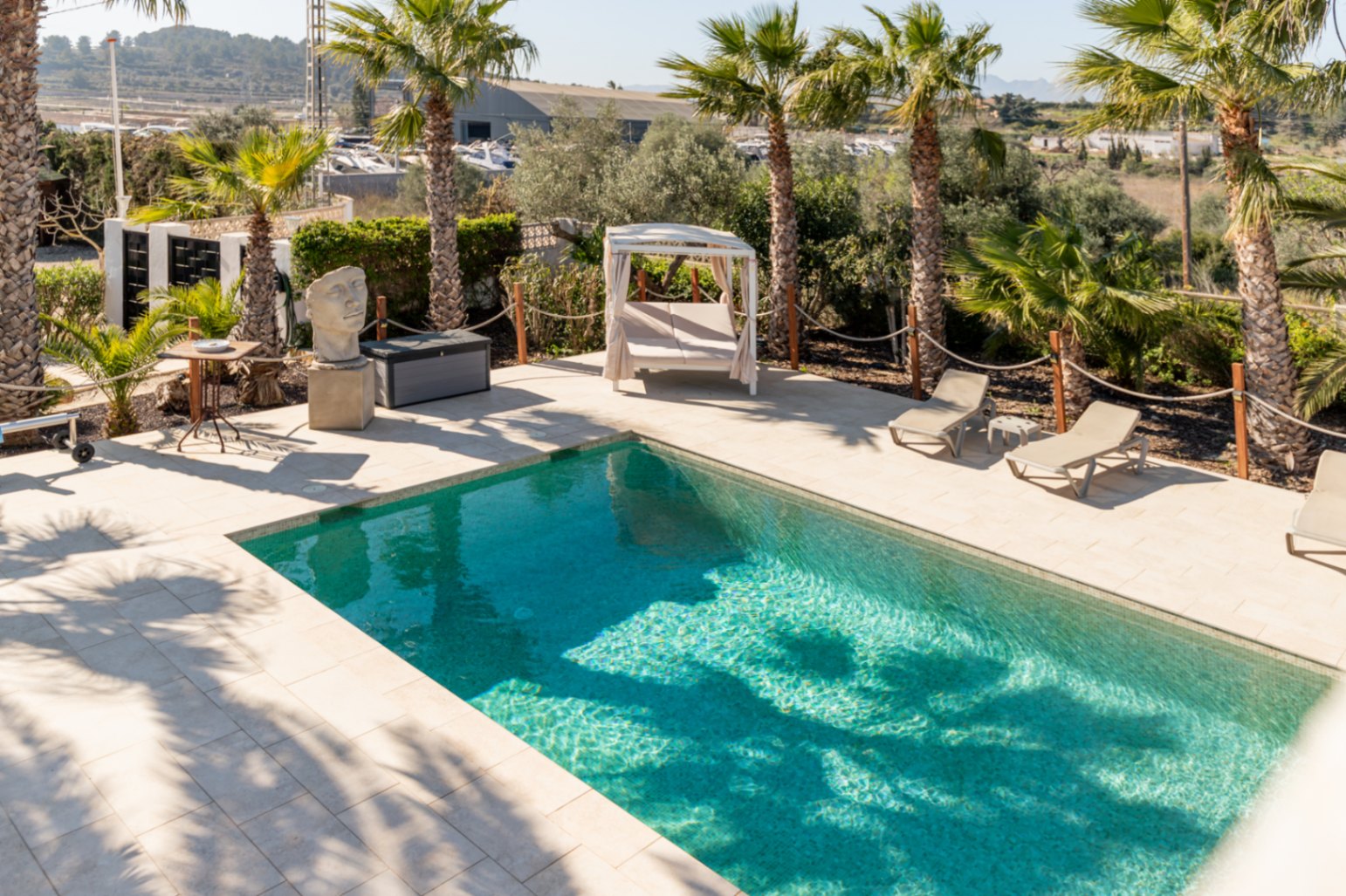Piscine rectangulaire avec terrasse en pierre claire, chaises longues et lit balinais, entourée de palmiers et d'une clôture en corde traditionnelle.