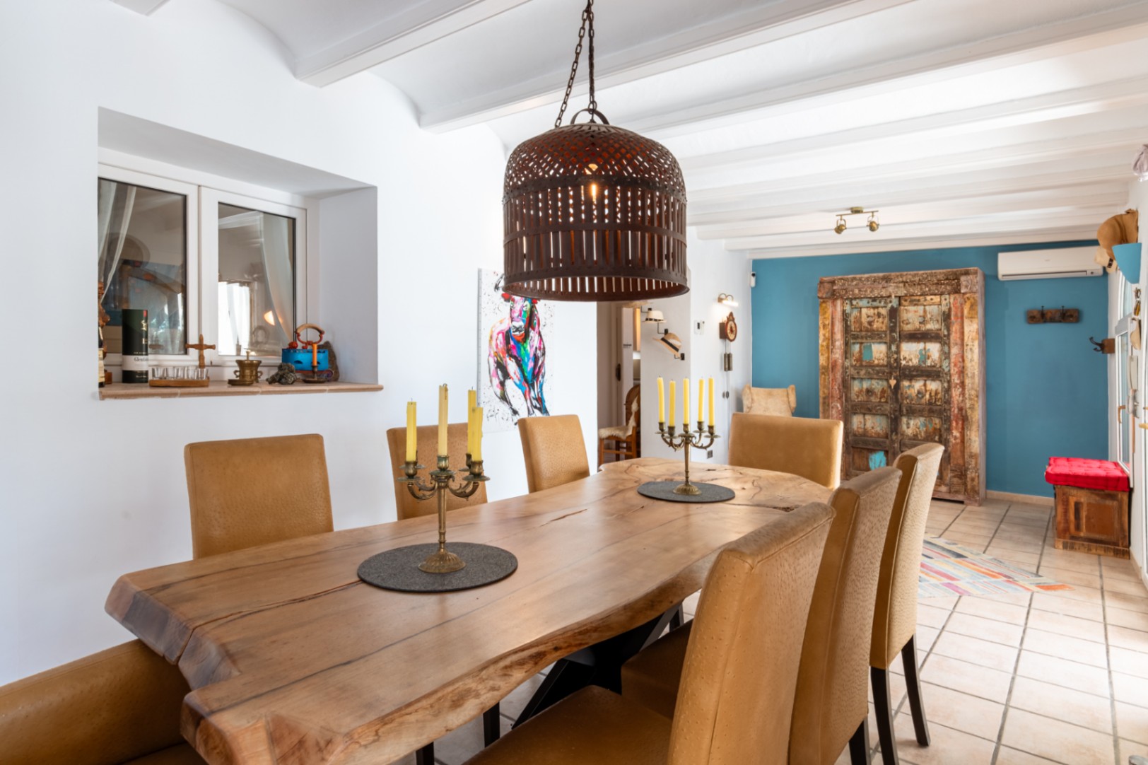 Salle à manger avec table en bois massif à bord naturel, chaises texturées et plafond à poutres blanches, soulignée par un mur d'accent bleu et des touches rustiques.