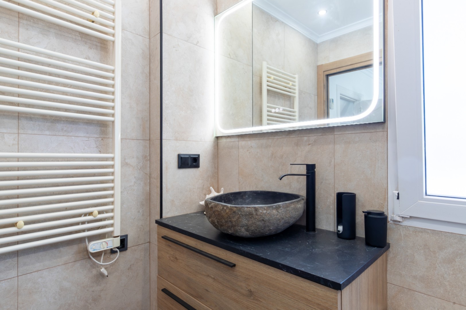 Salle de bain moderne avec vasque en pierre naturelle sur plan de travail noir et meuble en bois. Miroir rétroéclairé par LED et sèche-serviettes blanc mural.