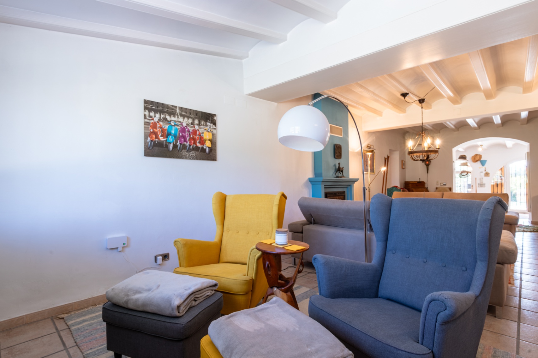Salon chaleureux avec plafonds à poutres blanches, cheminée bleue et fauteuils à oreilles colorés sous un lampadaire arc moderne.