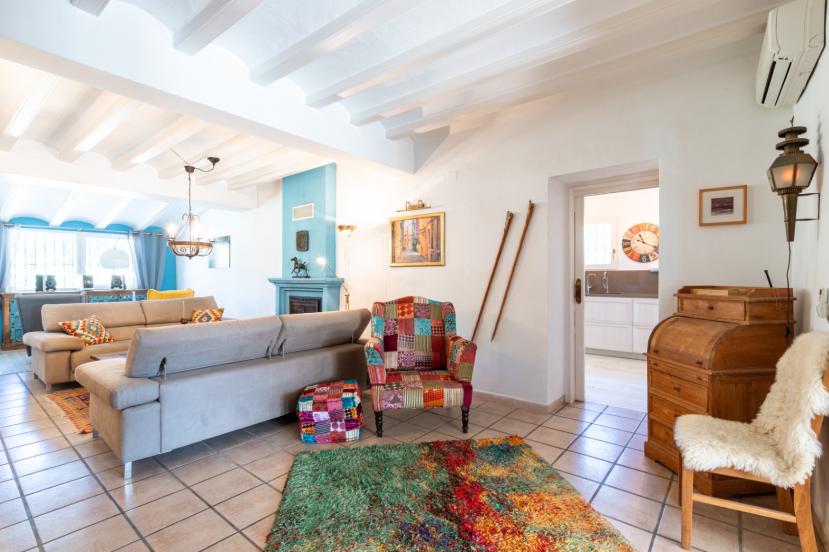 Salon lumineux avec plafonds à poutres blanches, cheminée bleue et mobilier éclectique. L'espace offre un grand canapé et une vue sur la cuisine attenante.