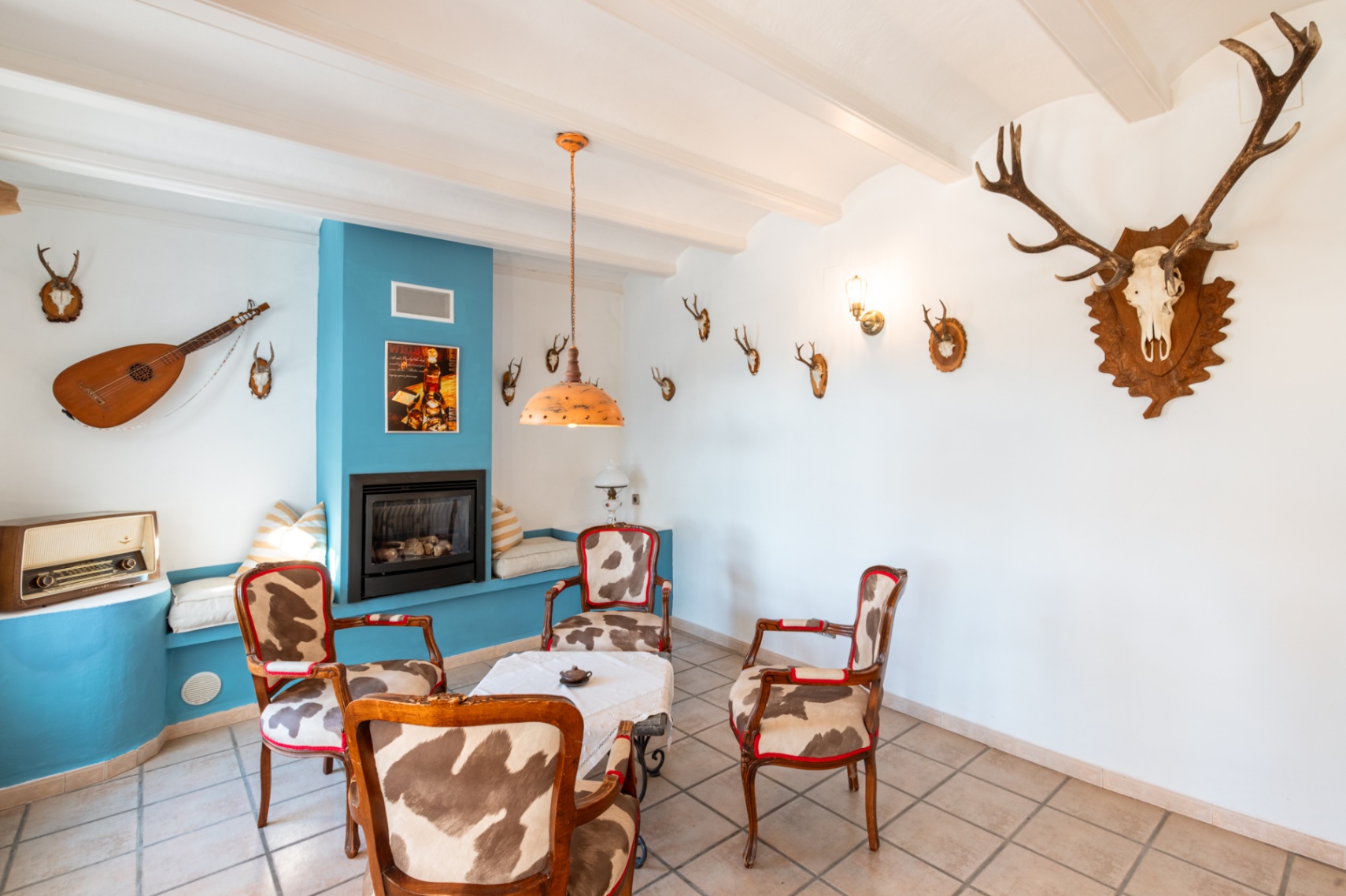 Salon rustique avec cheminée à gaz intégrée dans un mur bleu, fauteuils en peau de vache et plafond à poutres blanches orné de trophées de chasse.