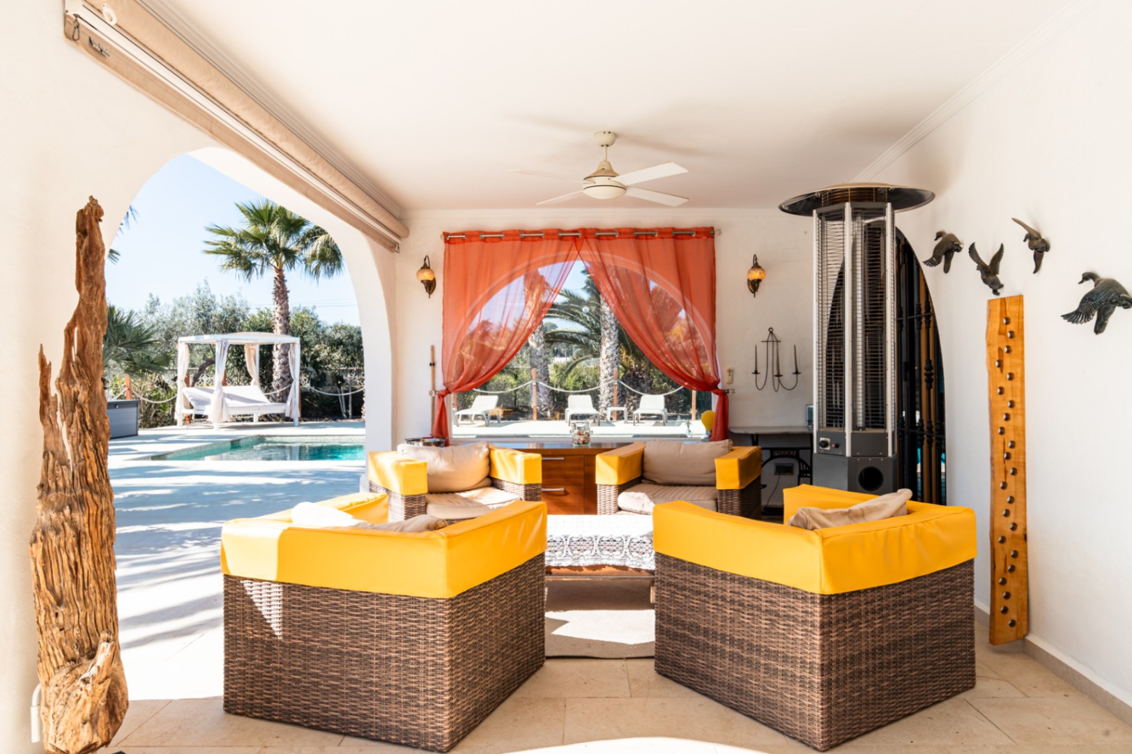 Terrasse couverte aménagée avec des fauteuils en osier, ventilateur de plafond et chauffage extérieur, offrant une vue dégagée sur la piscine et le jardin.