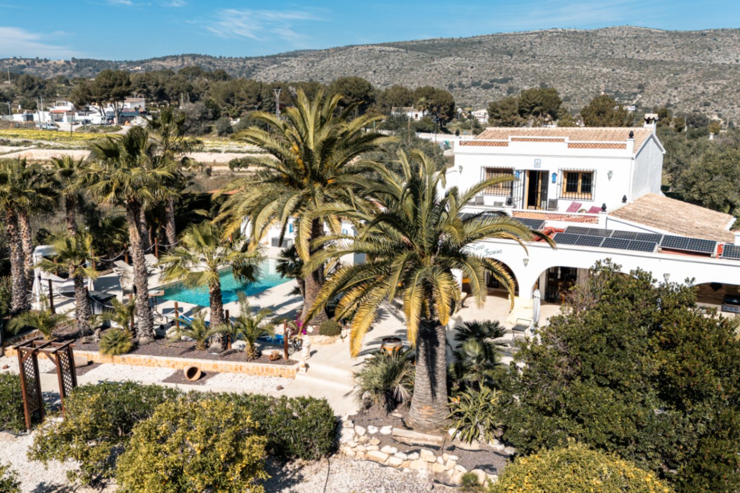 Vue aérienne de la propriété mettant en valeur les panneaux solaires, la piscine entourée de palmiers matures et l'architecture traditionnelle méditerranéenne.