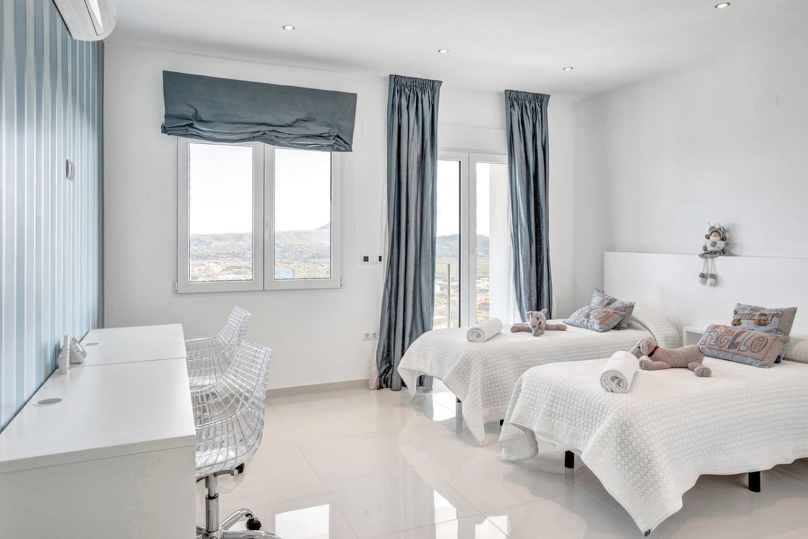 Chambre avec lits jumeaux, bureau blanc et chaises en polycarbonate, offrant une vue panoramique sur les montagnes et la mer Méditerranée.