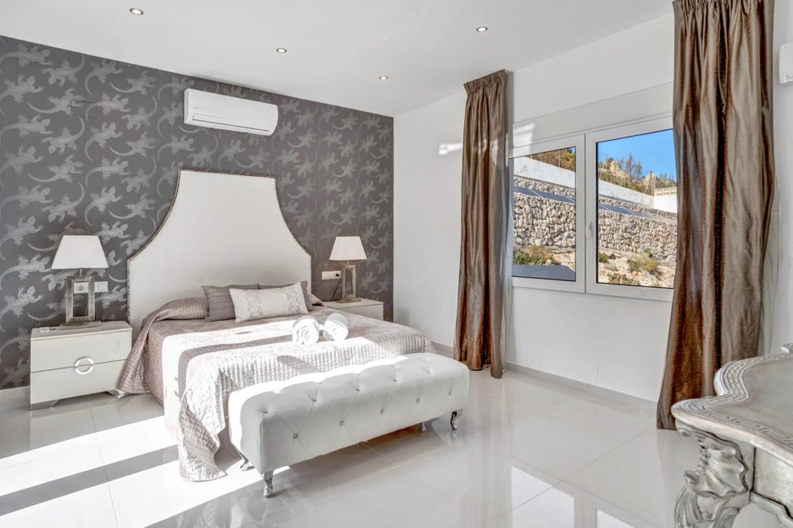 Chambre en suite avec mur d'accent gris à motifs, sol en carrelage blanc, climatisation et fenêtre offrant une vue sur le terrain rocheux environnant.