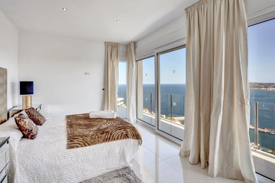 Chambre en suite avec sol en grès poli blanc, grandes baies vitrées et accès direct à un balcon offrant une vue panoramique sur la mer Méditerranée et le port de Jávea.