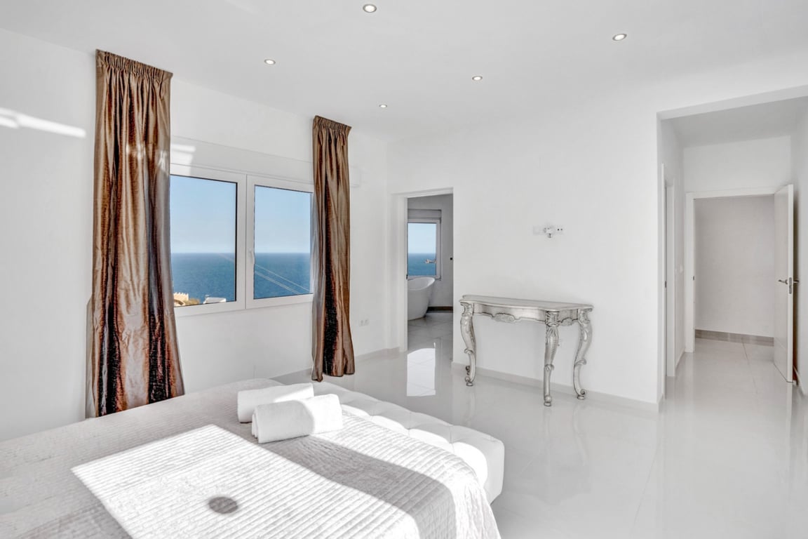 Chambre en suite avec sols en grès cérame blanc, vue sur mer et salle de bain intégrée disposant d'une baignoire îlot face à la Méditerranée.