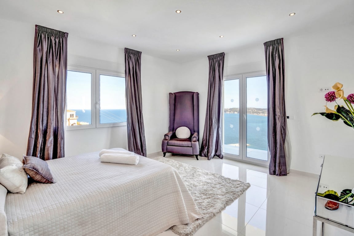 Chambre lumineuse avec fauteuil en velours violet, sol en carrelage blanc et grandes baies vitrées offrant une vue panoramique sur la mer.