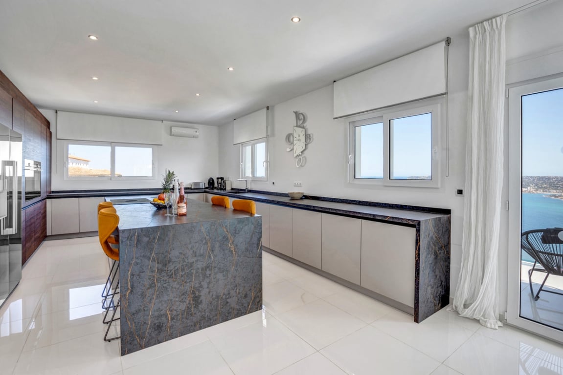 Cuisine contemporaine avec îlot en pierre sombre, tabourets moutarde et finitions épurées. Les larges baies vitrées offrent une vue dégagée sur la mer et la côte.