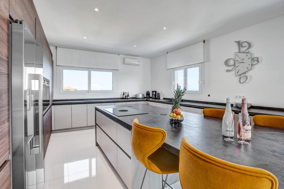 Cuisine design avec îlot central en pierre sombre, coin repas avec tabourets moutarde et rangements en bois et blanc sur un sol poli brillant.