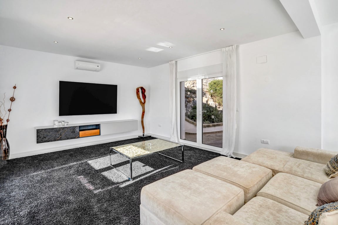 Espace multimédia moderne avec canapé modulaire, tapis épais et meuble suspendu, ouvrant sur une terrasse bordée par la roche naturelle de La Corona.