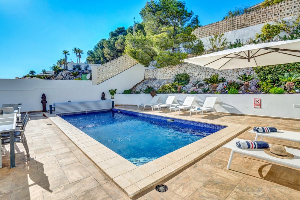 Piscine extérieure privée avec terrasse en pierre, bains de soleil blancs et parasol, bordée par un mur en pierre naturelle et une végétation locale.