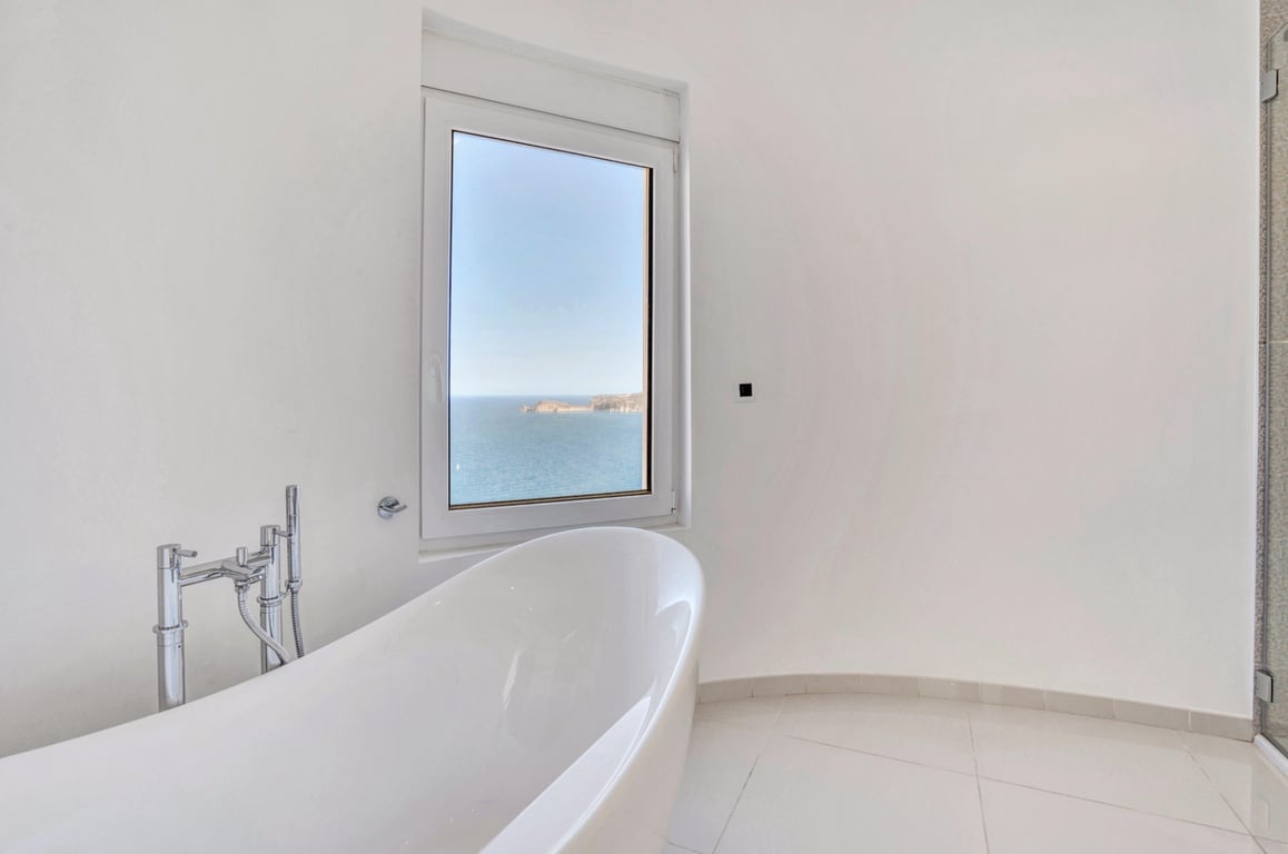 Salle de bain minimaliste avec baignoire îlot blanche et robinetterie chromée, offrant une vue dégagée sur la mer Méditerranée depuis la fenêtre.