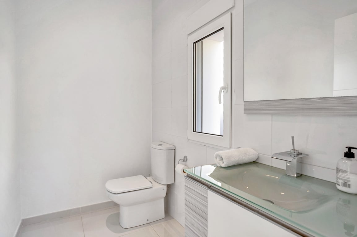 Salle de bain moderne avec plan de vasque en verre translucide, robinetterie chromée et murs carrelés blancs avec fenêtre.