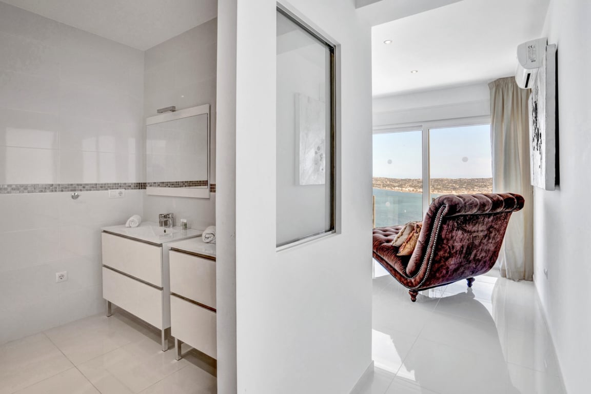 Salle de bains attenante avec double vasque et mosaïque, offrant une vue sur une chambre lumineuse équipée d'une chaise longue face à la mer.