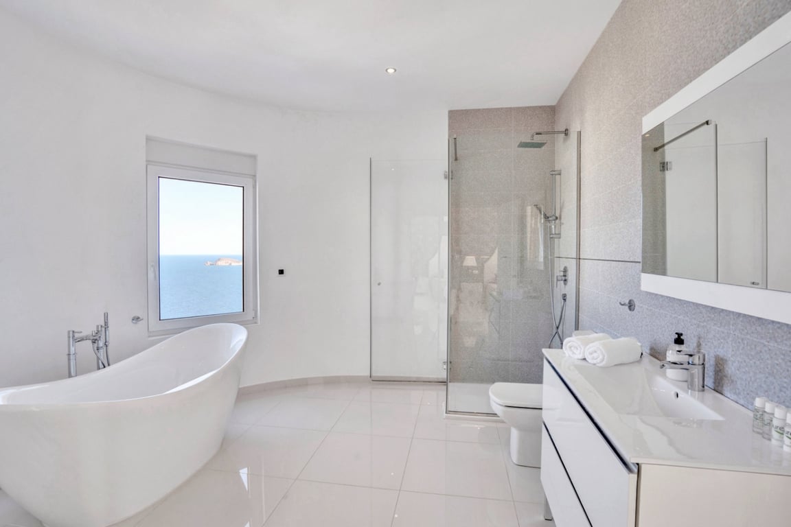 Salle de bains moderne avec baignoire îlot face à la mer, douche à l'italienne vitrée et meuble vasque blanc avec grand miroir intégré.