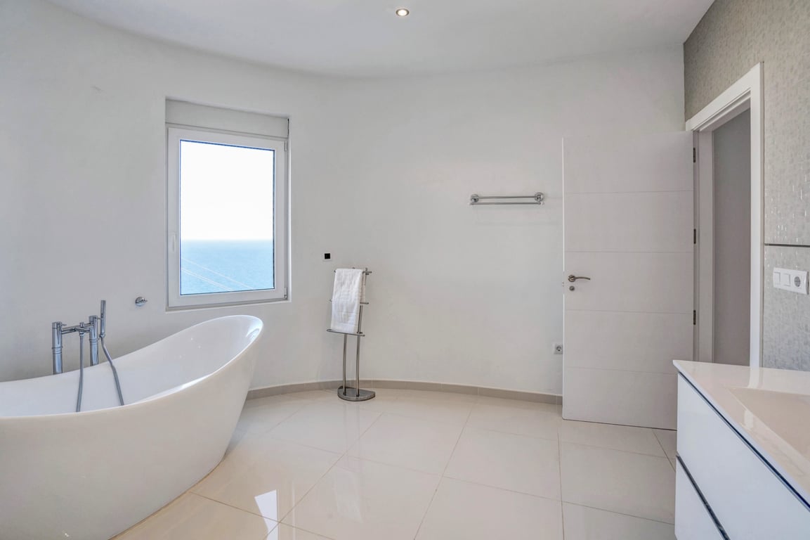 Salle de bains moderne avec baignoire îlot, sol en grès cérame clair et fenêtre offrant une vue dégagée sur la mer Méditerranée.