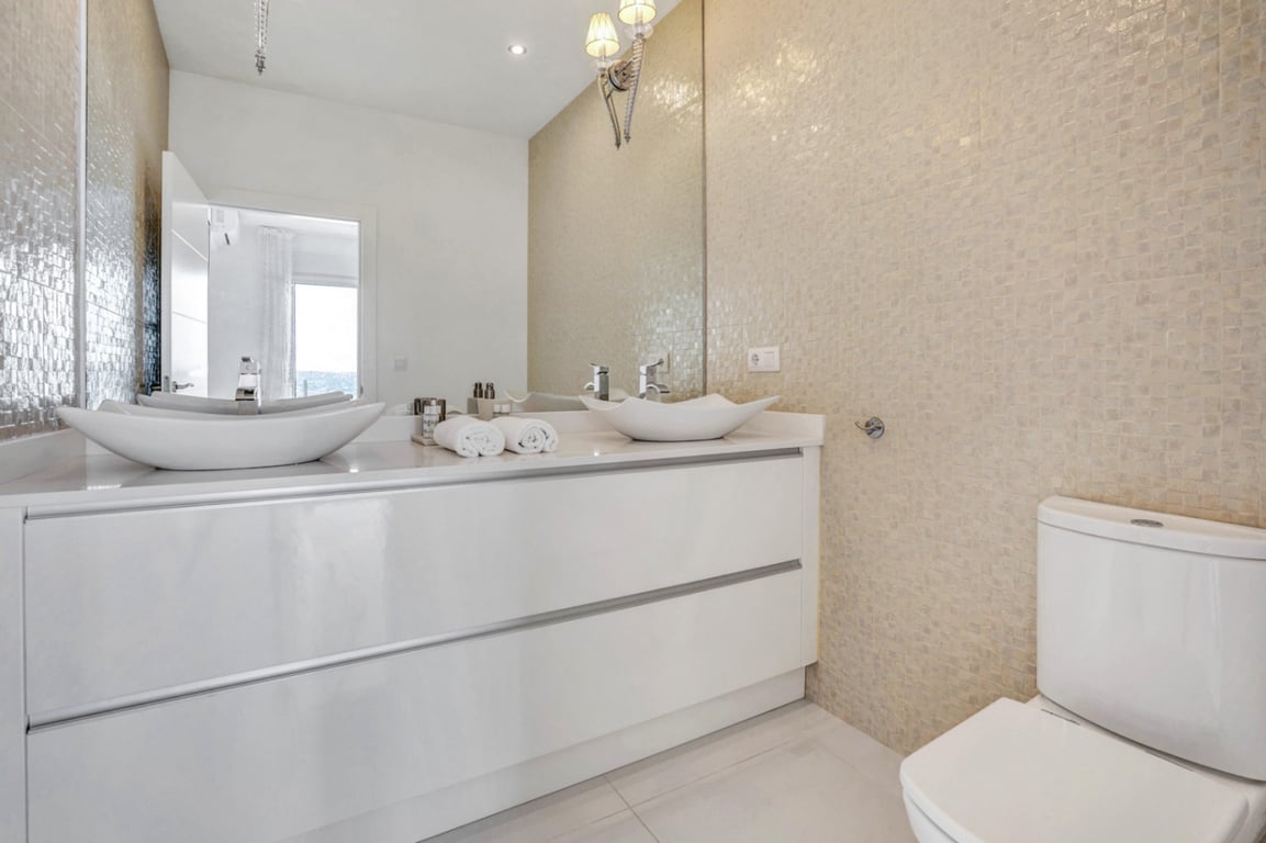Salle de bains moderne avec double vasque sur meuble blanc brillant, mur en mosaïque beige et grand miroir avec éclairage intégré.