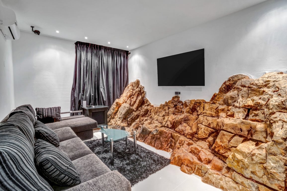 Salle de cinéma privée avec roche naturelle intégrée au mur, canapé gris, table basse en verre et télévision murale sous un éclairage encastré.