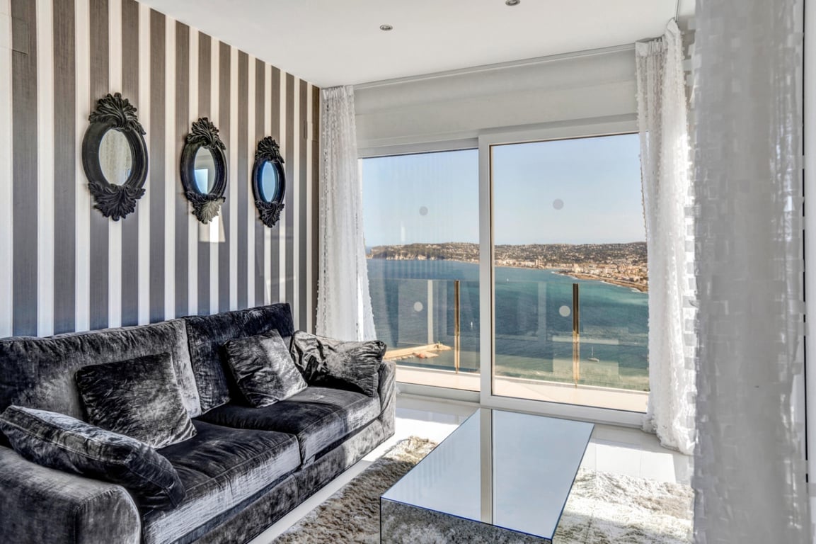 Salon élégant avec canapé en velours gris, papier peint à rayures et miroirs baroques, ouvrant sur une vue panoramique imprenable sur la baie de Jávea.