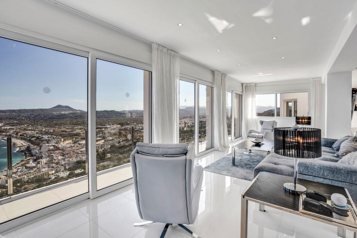 Salon lumineux doté de baies vitrées offrant une vue panoramique sur la baie de Jávea et les montagnes, avec un sol en carrelage blanc et un éclairage encastré.