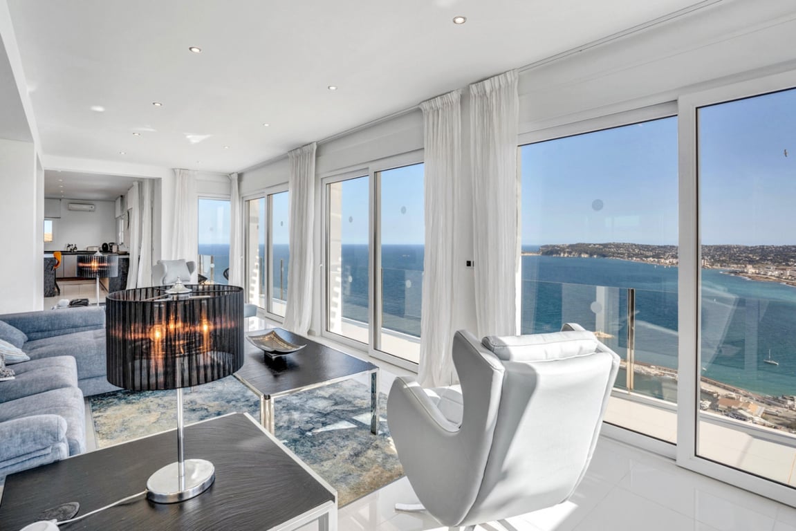 Séjour lumineux avec baies vitrées coulissantes offrant une vue panoramique sur la baie. L'espace dispose d'un mobilier moderne, de rideaux blancs et d'un sol en carrelage clair.