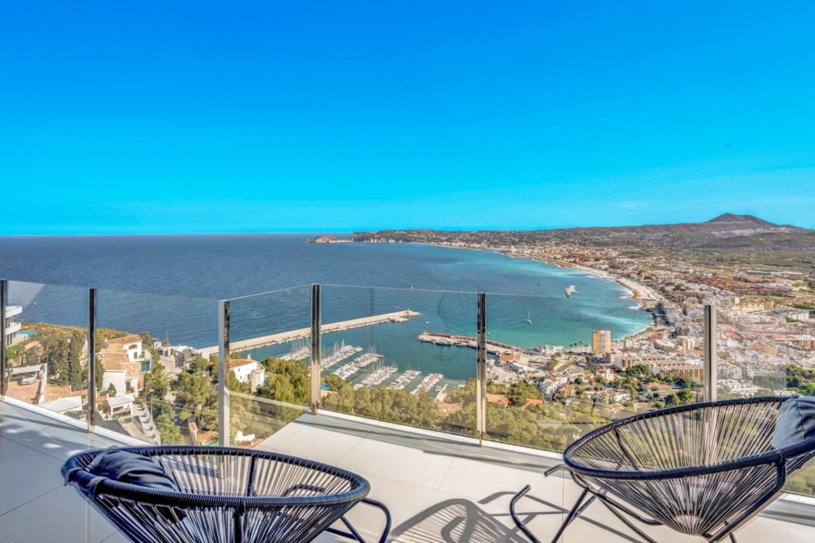 Terrasse avec garde-corps en verre et fauteuils Acapulco noirs, offrant une vue panoramique sur le port de Jávea et la côte méditerranéenne.