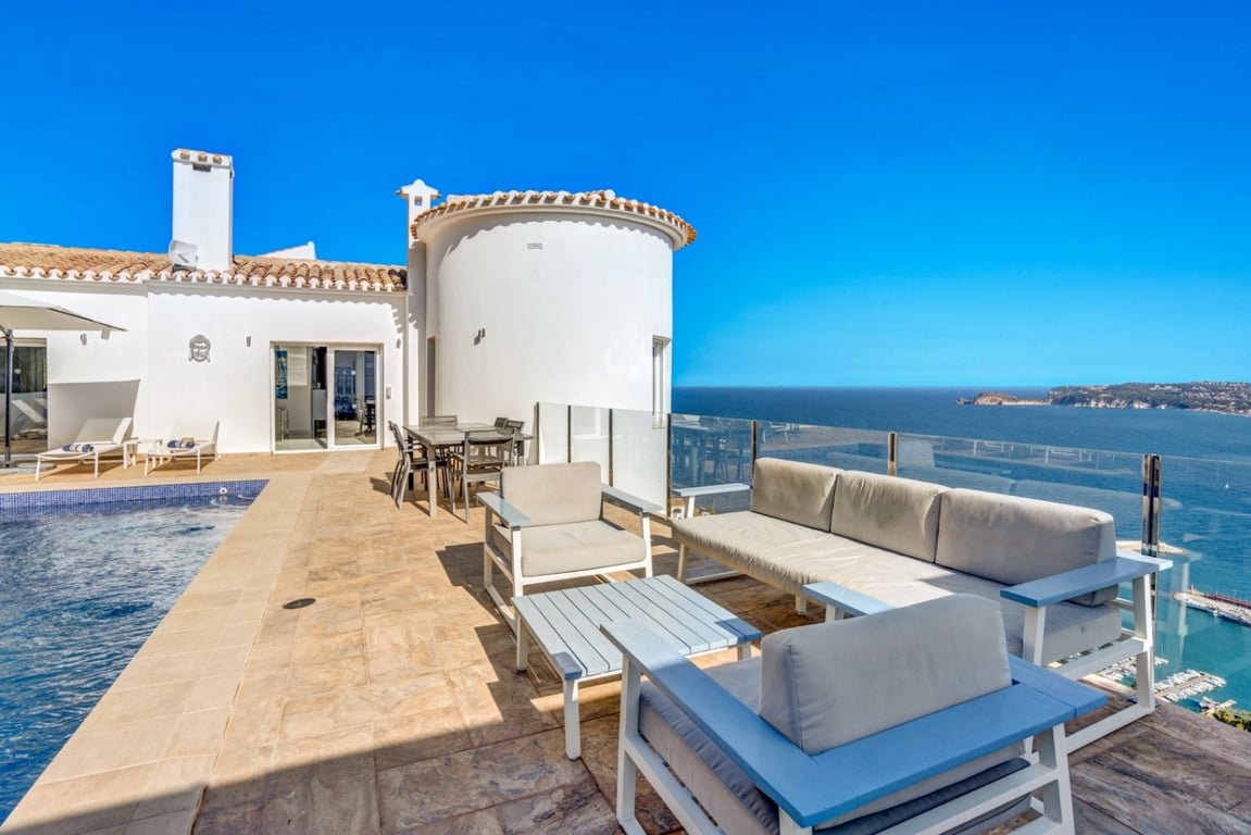 Terrasse principale avec salon d'été, espace dînatoire et piscine, offrant une vue panoramique sur le port de Jávea et la mer derrière un garde-corps en verre.