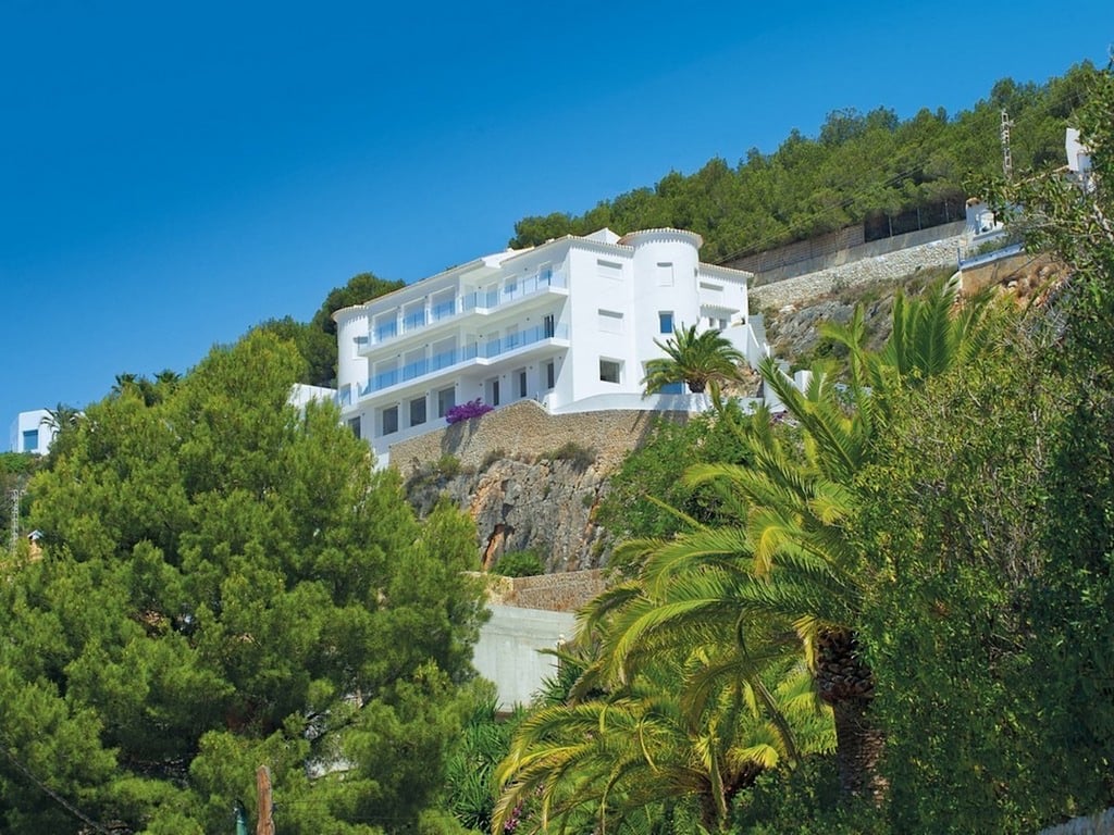 Vue extérieure de la villa blanche nichée dans la roche, offrant plusieurs niveaux de terrasses avec garde-corps en verre et une végétation méditerranéenne dense.