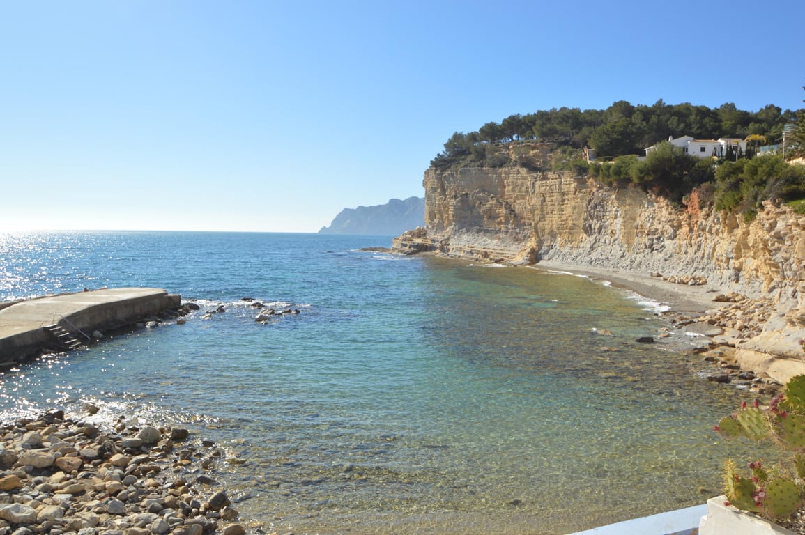 Vue sur la Cala Advocat avec ses eaux limpides, sa jetée en béton et ses falaises calcaires couronnées de pins parasols et de résidences côtières.