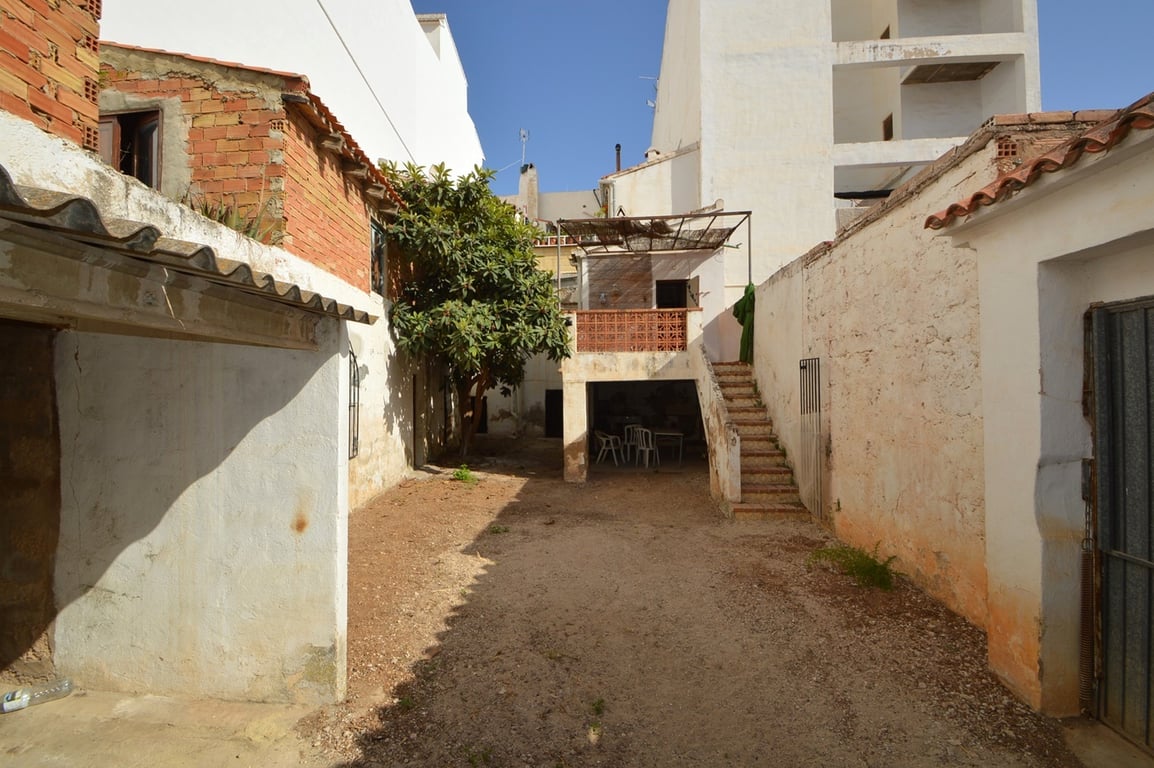 Cour intérieure privée avec sol en terre battue, hauts murs et escalier extérieur menant à une terrasse surélevée et un espace couvert sous auvent.