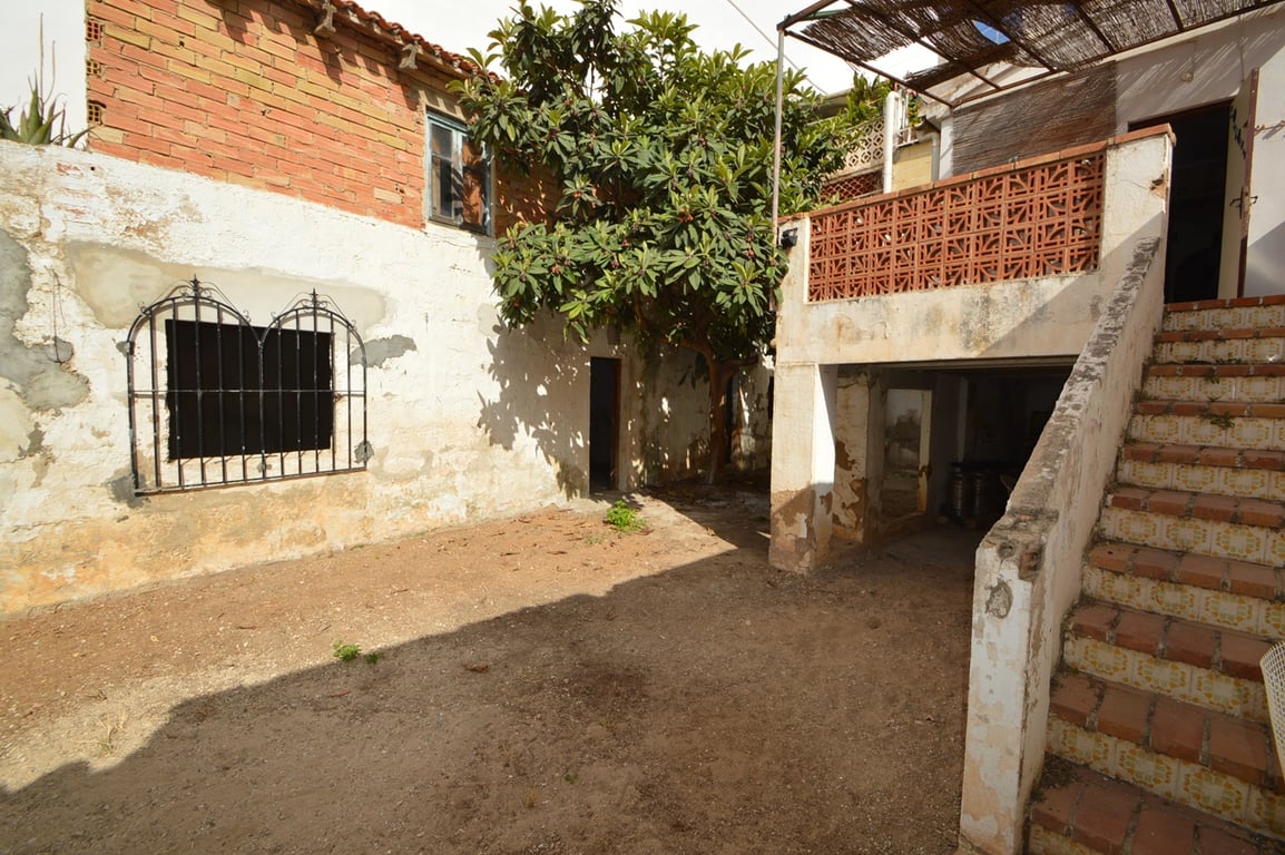 Cour intérieure rustique avec sol en terre, grand néflier et escalier extérieur carrelé menant à une terrasse avec pergola en canisse et garde-corps en céramique.