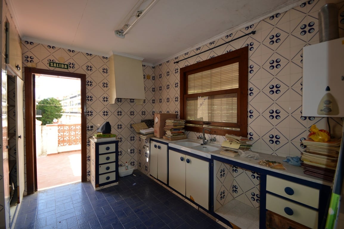 Cuisine avec carrelage mural à motifs, sol bleu et accès direct à la terrasse. Elle comprend un double évier, une fenêtre en bois et des meubles aux finitions bleues.
