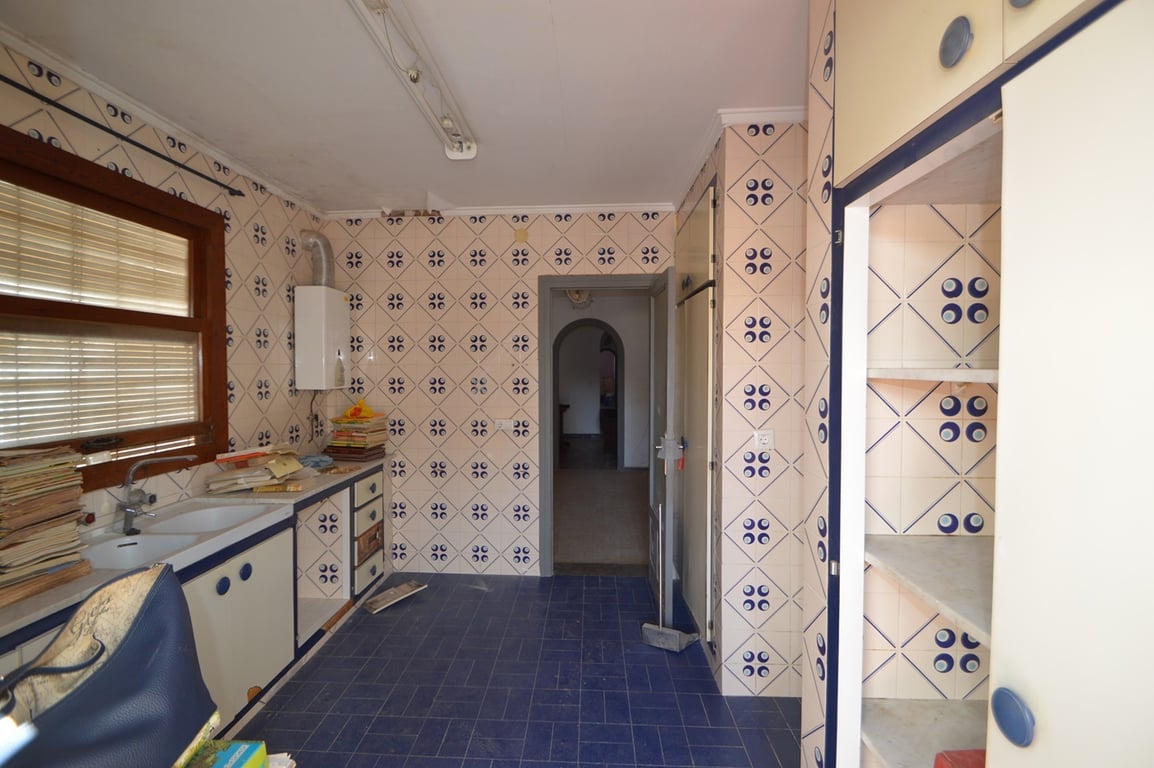 Cuisine avec carrelage mural décoratif, sol bleu et placards blancs aux finitions bleues. Comprend un double évier sous une fenêtre en bois et mène vers un couloir intérieur.