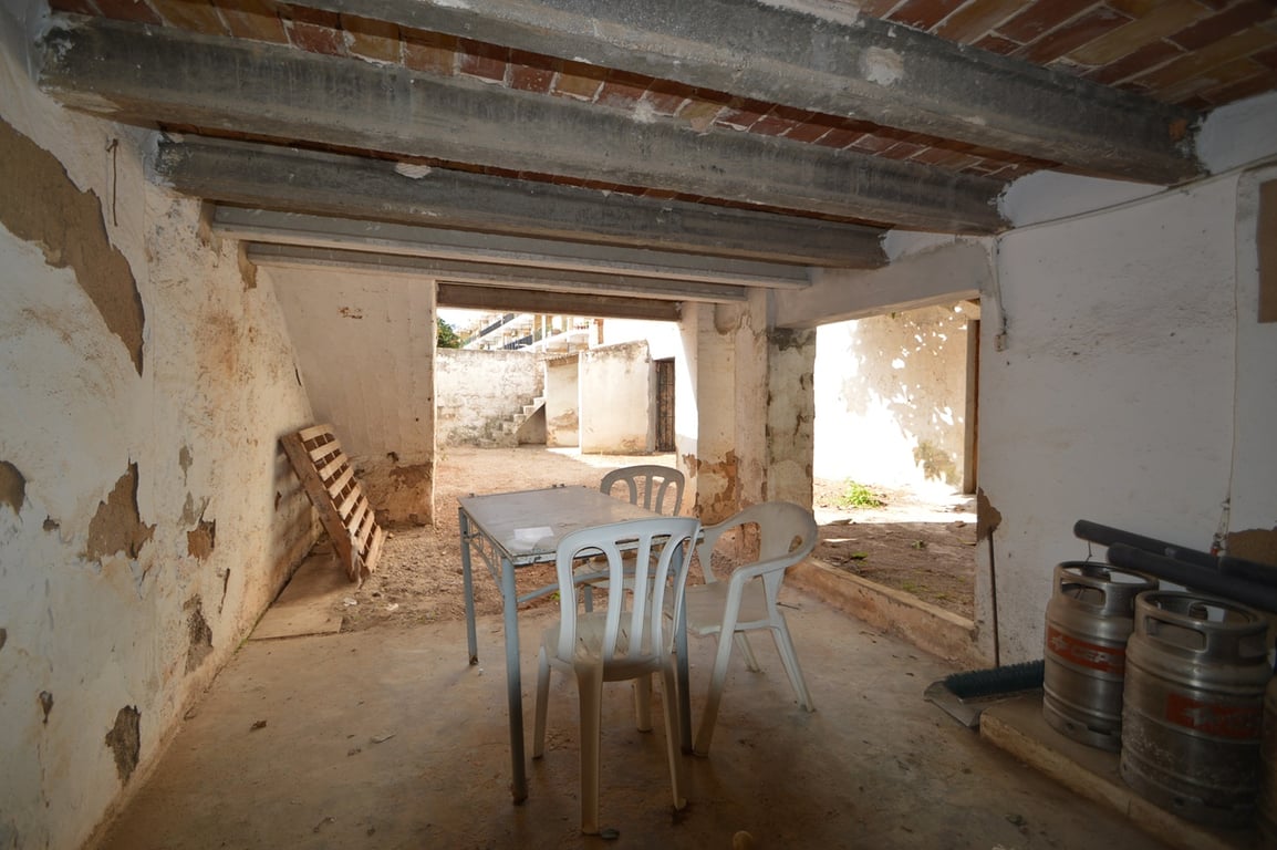 Espace couvert avec poutres en béton apparentes et briques, s'ouvrant sur une cour intérieure privée et un jardin clos.
