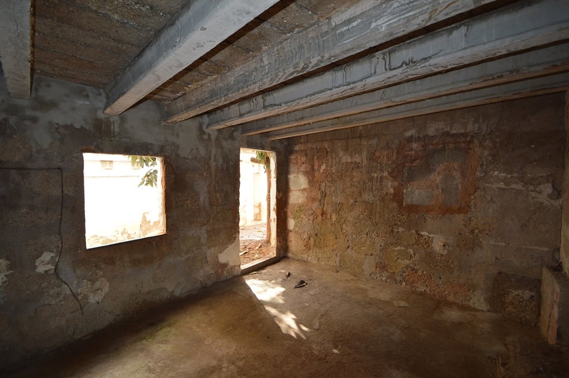 Espace intérieur brut avec murs en pierre d'origine et poutres apparentes au plafond. La lumière naturelle pénètre par une fenêtre et une porte menant à la cour.