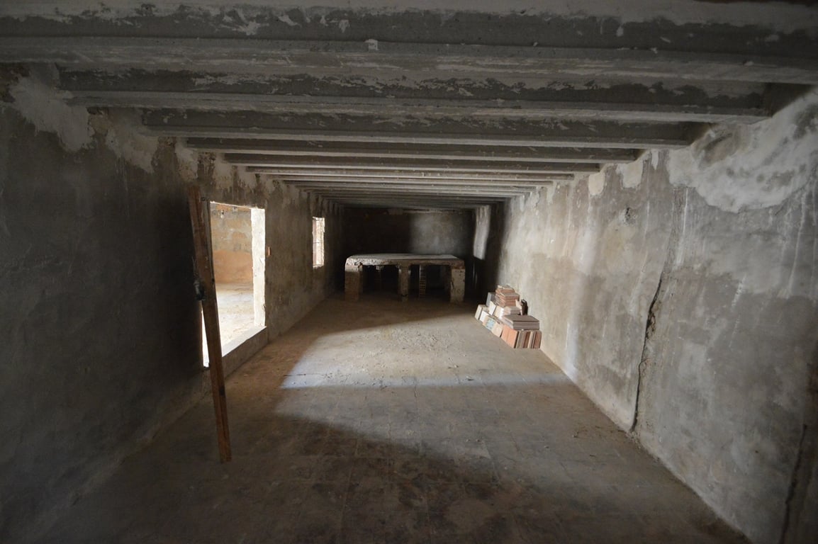 Grand espace brut avec poutres en béton apparentes, murs en pierre et une banquette maçonnée, idéal pour un atelier ou une zone de stockage.