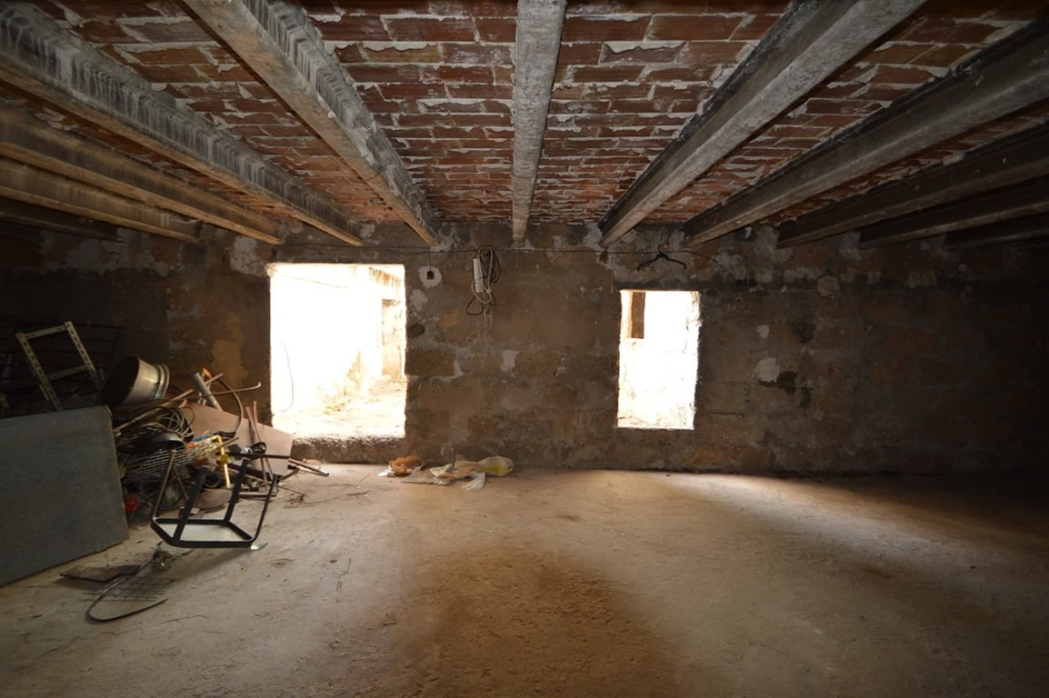 Intérieur spacieux à rénover avec plafonds en poutres de béton et briques apparentes. La pièce dispose de murs en pierre naturelle et de deux ouvertures vers l'extérieur.